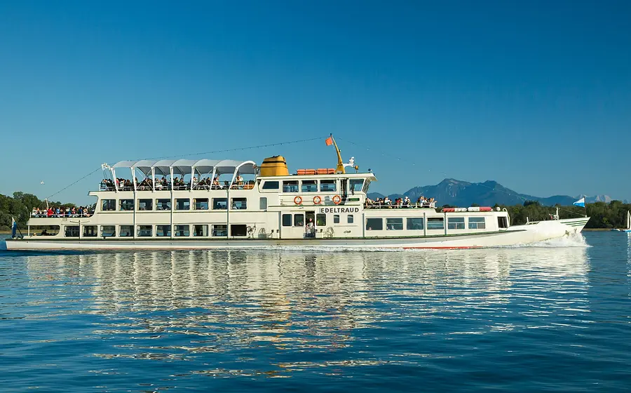 Fahrgastschiff Edeltraud mit mehreren Passagieren auf den Außendecks, unterwegs auf einem See vor Bergkulisse und blauem Himmel, mit Spiegelung des Schiffes im ruhigen Wasser