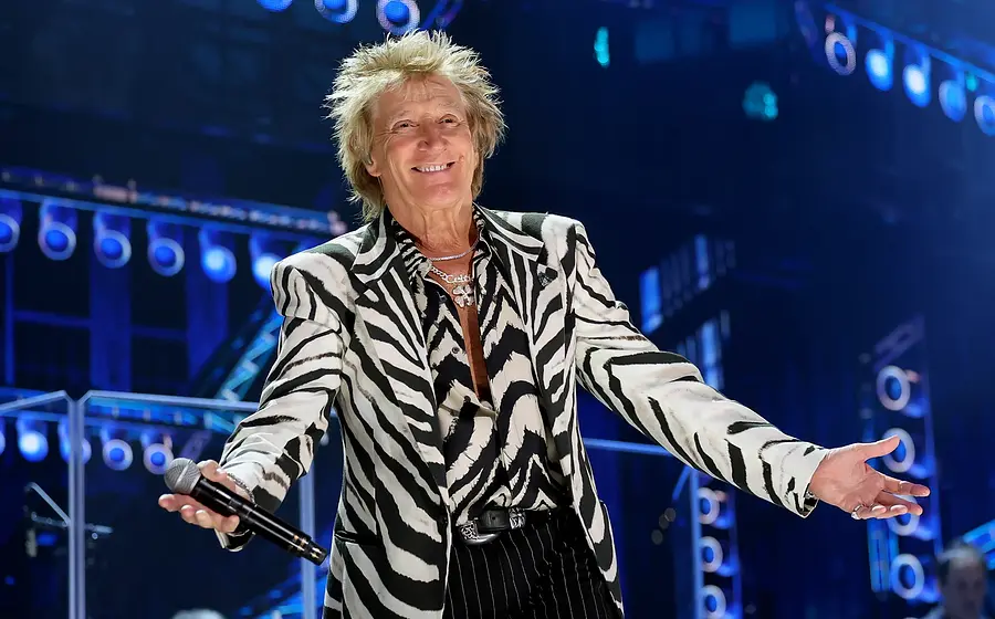 Bühnenfoto von Rod Stewart in schwarz weiß gemustertem Sakko mit Mikrofon in der Hand auf einer blau beleuchteten Konzertbühne, umgeben von Scheinwerfern und Bühnenelementen im Hintergrund