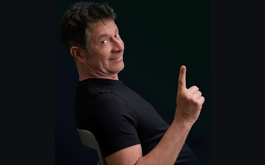 Rolf Miller auf einem Stuhl in schwarzem T-Shirt und erhobenem Zeigefinger blickt seitlich in die Kamera