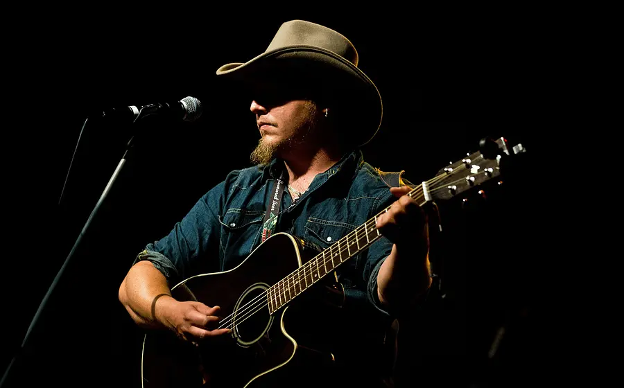 Der Künstler Roman Hofbauer mit Cowboyhut auf spielt Akustikgitarre auf einer dunklen Bühne vor einem Mikrofon und wird von einem einzelnen Scheinwerfer beleuchtet.