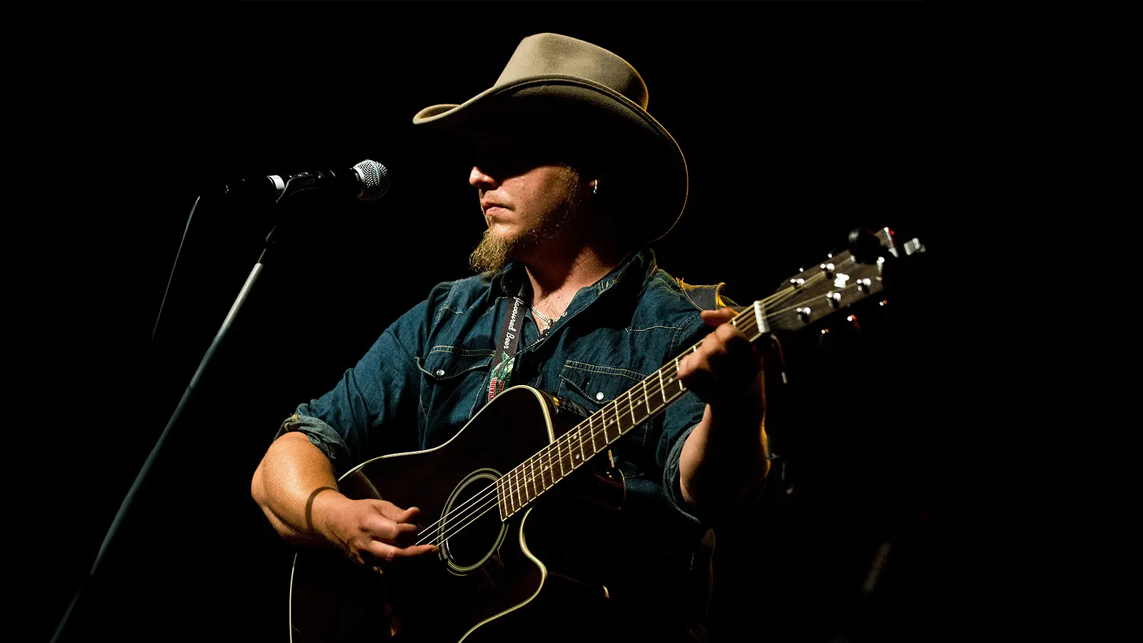 Der Künstler Roman Hofbauer mit Cowboyhut auf spielt Akustikgitarre auf einer dunklen Bühne vor einem Mikrofon und wird von einem einzelnen Scheinwerfer beleuchtet.