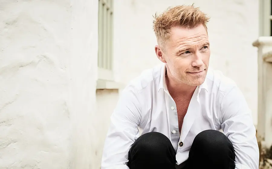 Ronan Keating in weißem Hemd und schwarzer Hose, sitzend vor heller Hausfassade mit Fenster und Balustrade, den Blick zur Seite gerichtet