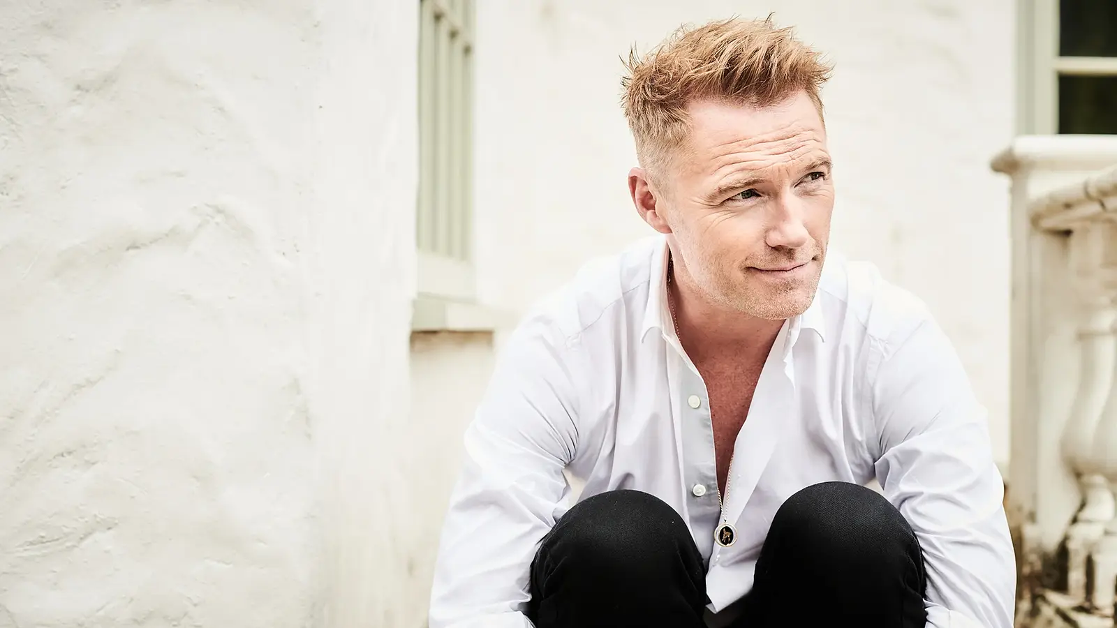 Ronan Keating in weißem Hemd und schwarzer Hose, sitzend vor heller Hausfassade mit Fenster und Balustrade, den Blick zur Seite gerichtet