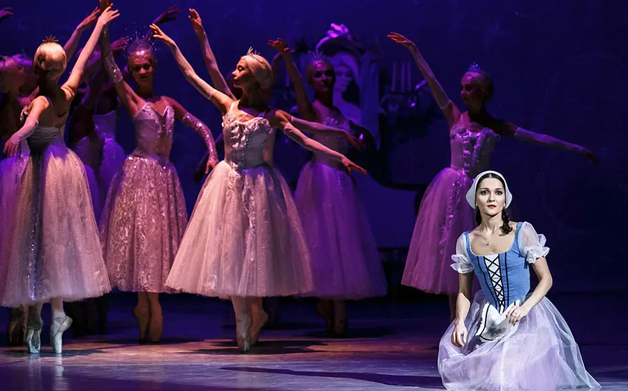 Bühnenfoto aus Cinderella mit mehreren Balletttänzerinnen in weißen Tüllkleidern und Diademen bei einer Tanzszene unter violettem Bühnenlicht, im Vordergrund eine Darstellerin in blau weißem Kleid kniend auf der Bühne.