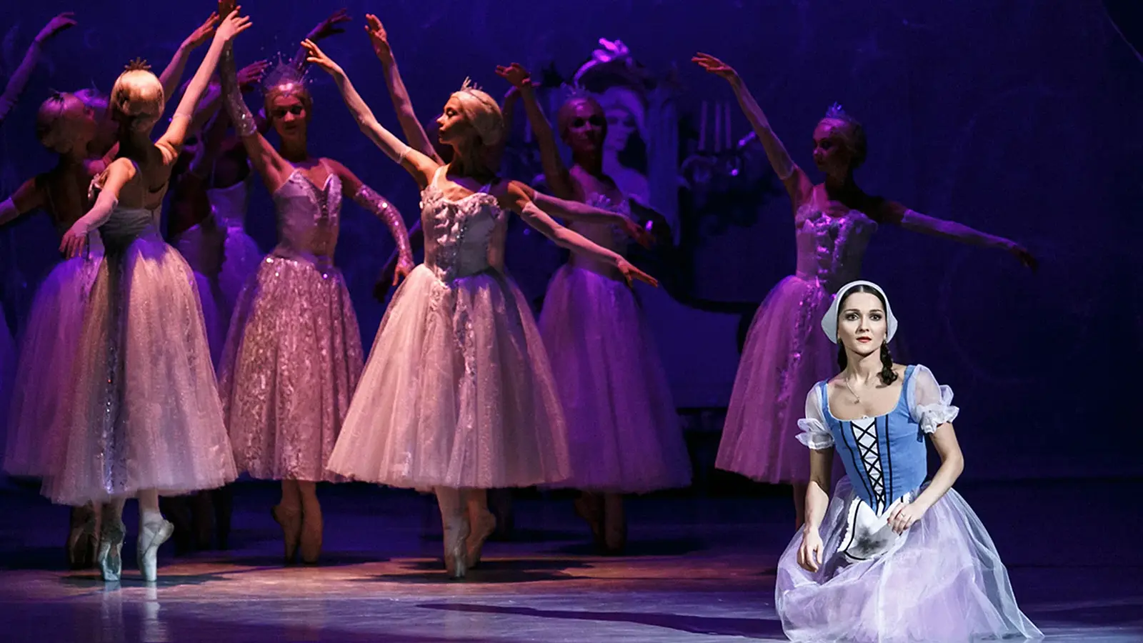 Bühnenfoto aus Cinderella mit mehreren Balletttänzerinnen in weißen Tüllkleidern und Diademen bei einer Tanzszene unter violettem Bühnenlicht, im Vordergrund eine Darstellerin in blau weißem Kleid kniend auf der Bühne.
