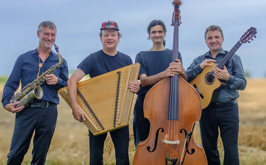 Vier Personen der Gruppe Rudi Zapf & Zapf’nstreich stehen auf einem Feld und posieren mit Saxofon, Hackbrett, Kontrabass und Gitarre vor blauem Himmel