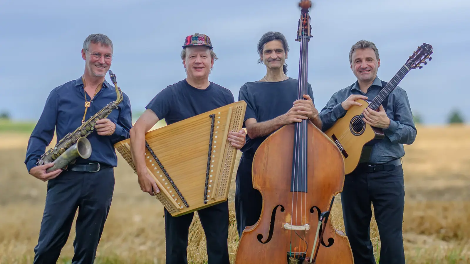 Vier Personen der Gruppe Rudi Zapf & Zapf’nstreich stehen auf einem Feld und posieren mit Saxofon, Hackbrett, Kontrabass und Gitarre vor blauem Himmel