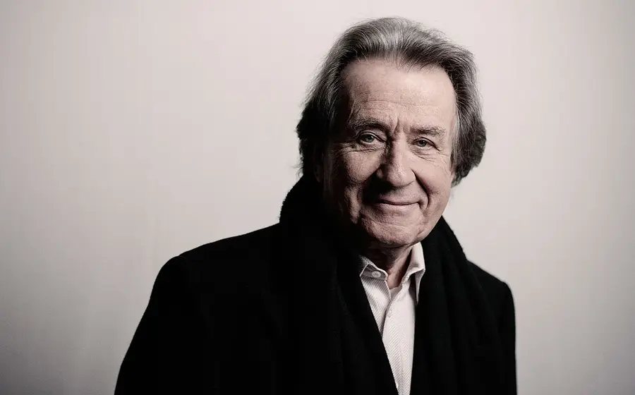 Portrait von Rudolf Buchbinder, lächelnd in hellem Hemd und schwarzem Mantel vor hellgrauem Hintergrund