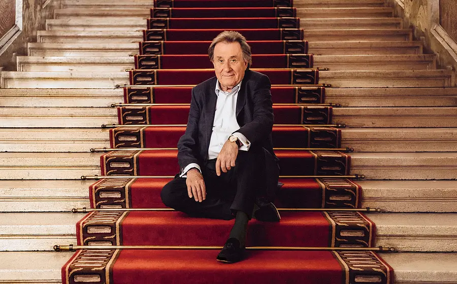 Rudolf Buchbinder in dunklem Anzug sitzt auf breiter, hellsteinener Treppe mit rotem Teppichläufer und goldenen Stangen in einem repräsentativen Innenraum