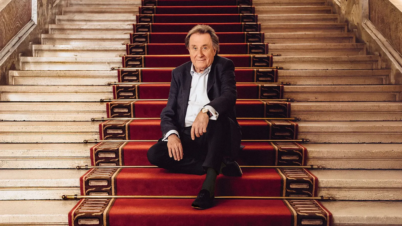 Rudolf Buchbinder in dunklem Anzug sitzt auf breiter, hellsteinener Treppe mit rotem Teppichläufer und goldenen Stangen in einem repräsentativen Innenraum