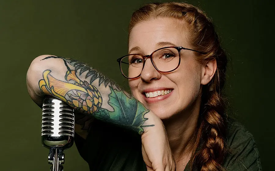 Porträtfoto von Sara Brandhuber mit Brille und langem geflochtenem Zopf, die sich auf ein Retro-Mikrofon stützt, vor grünem Hintergrund, mit auffälligem Tattoo am Unterarm.