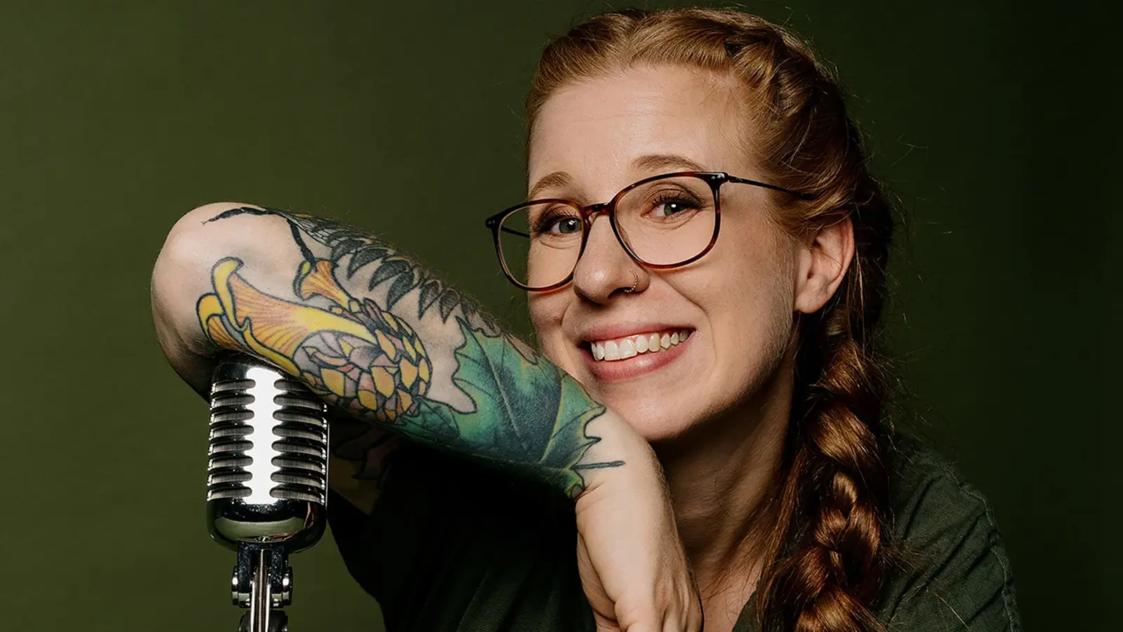 Porträtfoto von Sara Brandhuber mit Brille und langem geflochtenem Zopf, die sich auf ein Retro-Mikrofon stützt, vor grünem Hintergrund, mit auffälligem Tattoo am Unterarm.