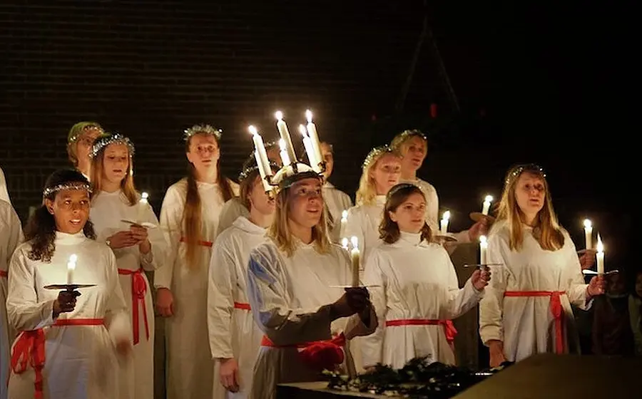 Luciachor mit Kerzen in weißen Gewändern bei einem Lichterzug. Die Personen tragen rote Gürtel und Kerzenkränze auf dem Kopf und halten brennende Kerzen in den Händen.