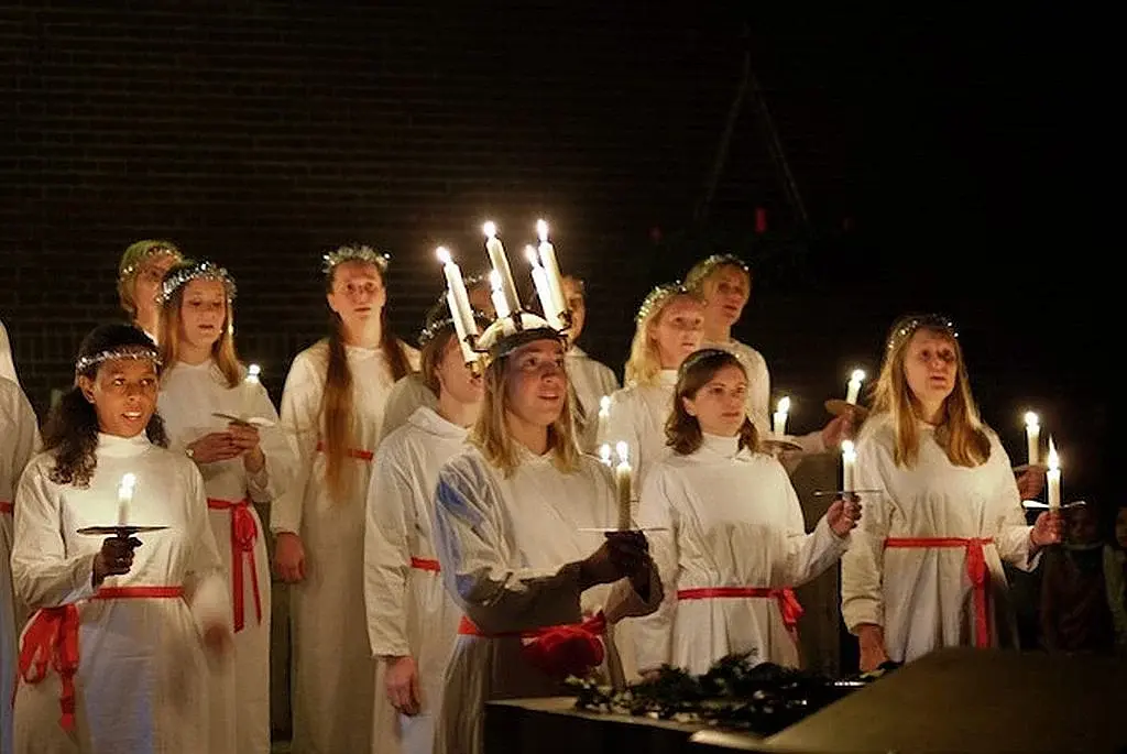 Luciachor mit Kerzen in weißen Gewändern bei einem Lichterzug. Die Personen tragen rote Gürtel und Kerzenkränze auf dem Kopf und halten brennende Kerzen in den Händen.