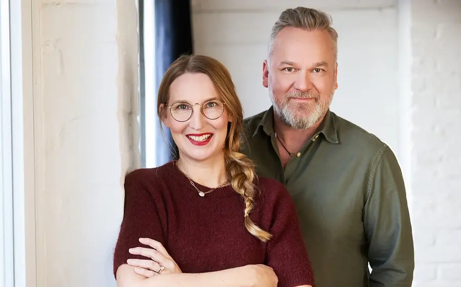 Franca Cerutti in dunkelrotem Pulli und Christian Weiss in grünem Hemd posieren vor einer weißen Mauer