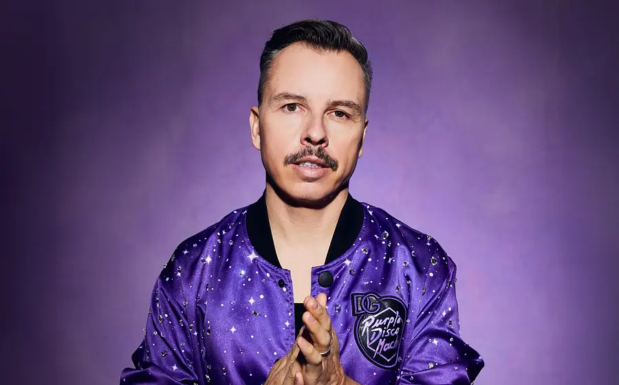Purple Disco Machine in einer violetten Jacke mit Glitzerapplikationen blickt direkt in die Kamera, vor violettem Hintergrund