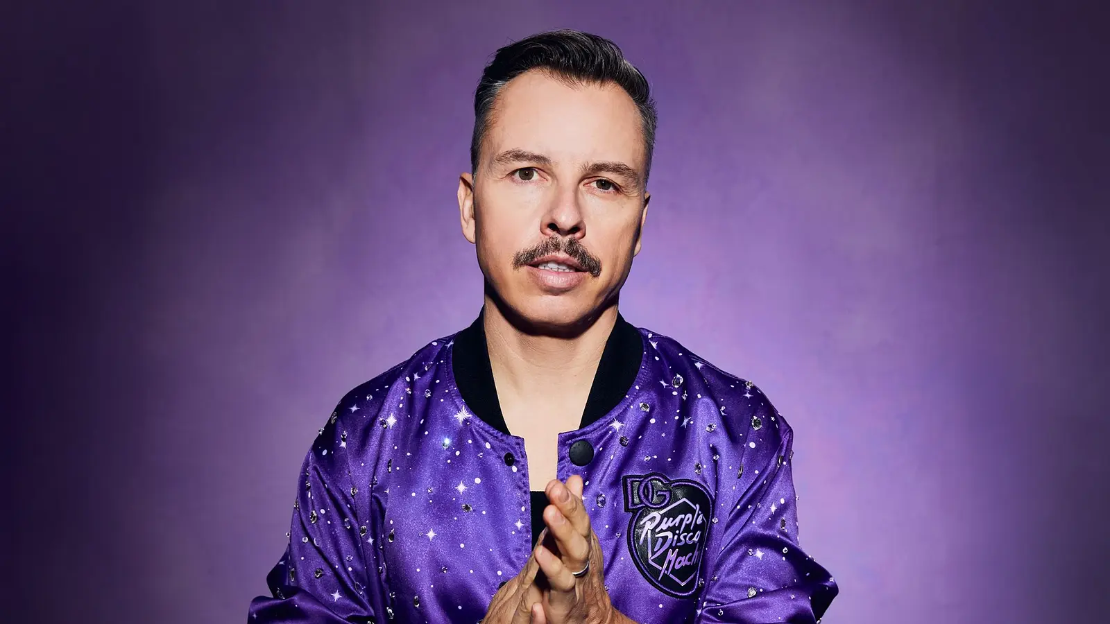 Purple Disco Machine in einer violetten Jacke mit Glitzerapplikationen blickt direkt in die Kamera, vor violettem Hintergrund