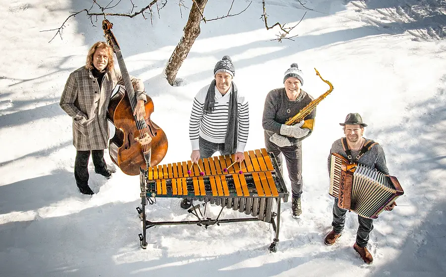 Vier Musiker on Quadro Nuevo mit Kontrabass, Vibraphon, Saxofon und Akkordeon stehen im Schnee unter einem Baum. Geben Sie Ihr Feedback zu BizChat ab.