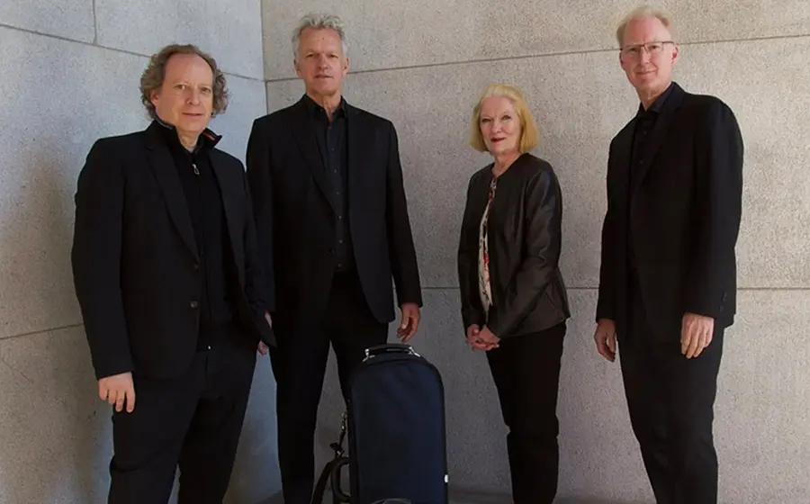 Personengruppe des American String Quartet vor einer hellen Steinwand. Die vier Ensemblemitglieder tragen dunkle Konzertkleidung, zwischen ihnen steht ein Instrumentenkoffer
