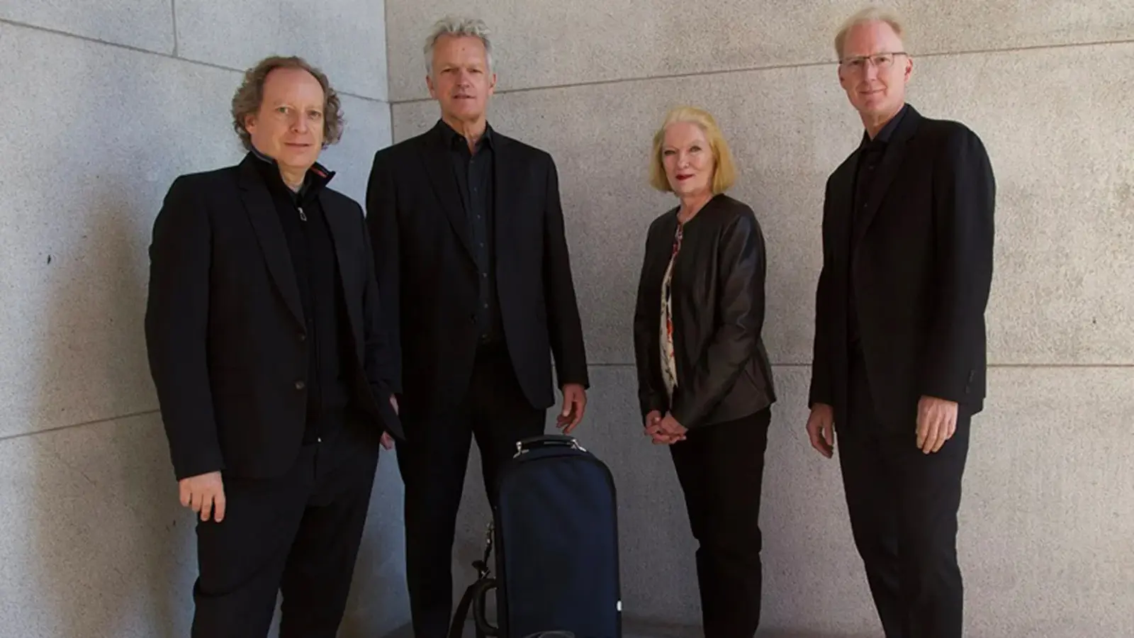 Personengruppe des American String Quartet vor einer hellen Steinwand. Die vier Ensemblemitglieder tragen dunkle Konzertkleidung, zwischen ihnen steht ein Instrumentenkoffer