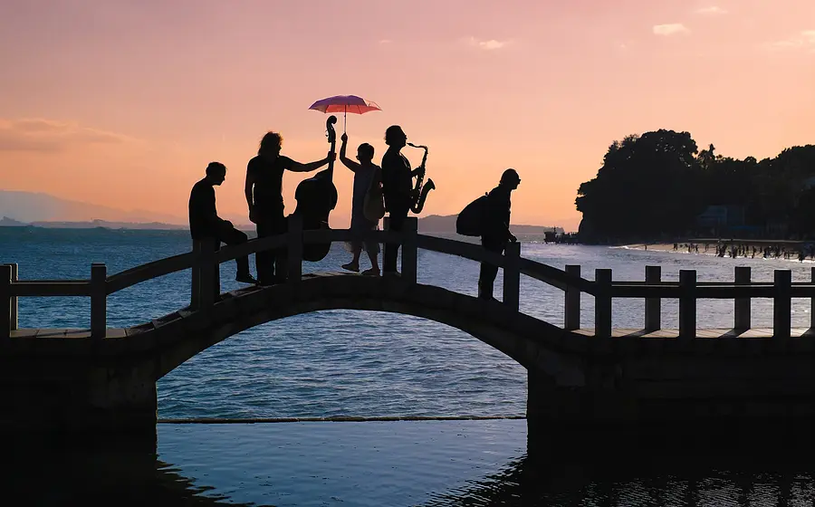 Silhouetten von fünf Mitgliedern von Quadro Nuevo auf einer geschwungenen Brücke über dem Wasser bei Sonnenuntergang, mit Kontrabass, Saxofon und einem aufgespannten Schirm vor Küstenlandschaft und Bergkulisse