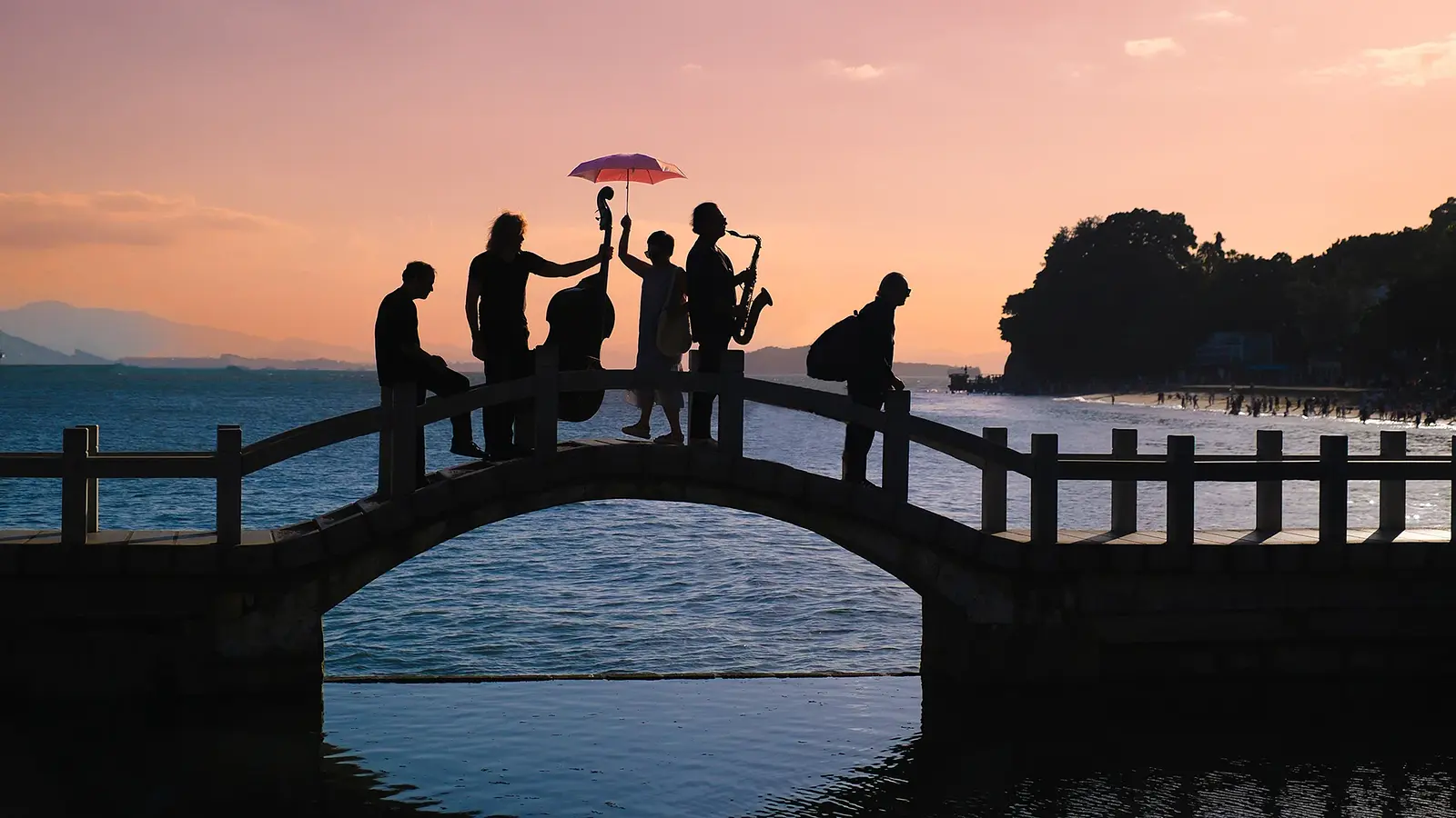 Silhouetten von fünf Mitgliedern von Quadro Nuevo auf einer geschwungenen Brücke über dem Wasser bei Sonnenuntergang, mit Kontrabass, Saxofon und einem aufgespannten Schirm vor Küstenlandschaft und Bergkulisse
