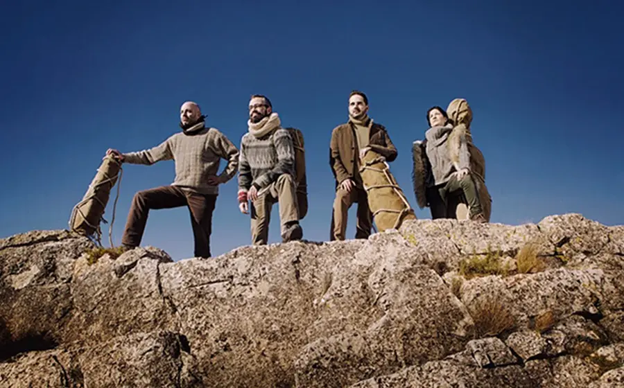 Personengruppe auf einer felsigen Anhöhe mit Instrumentenkoffern vor klarem blauem Himmel. Die Mitglieder des Quiroga Quartetts stehen in Outdoor-Kleidung auf einem Felsrücken und blicken in die Ferne