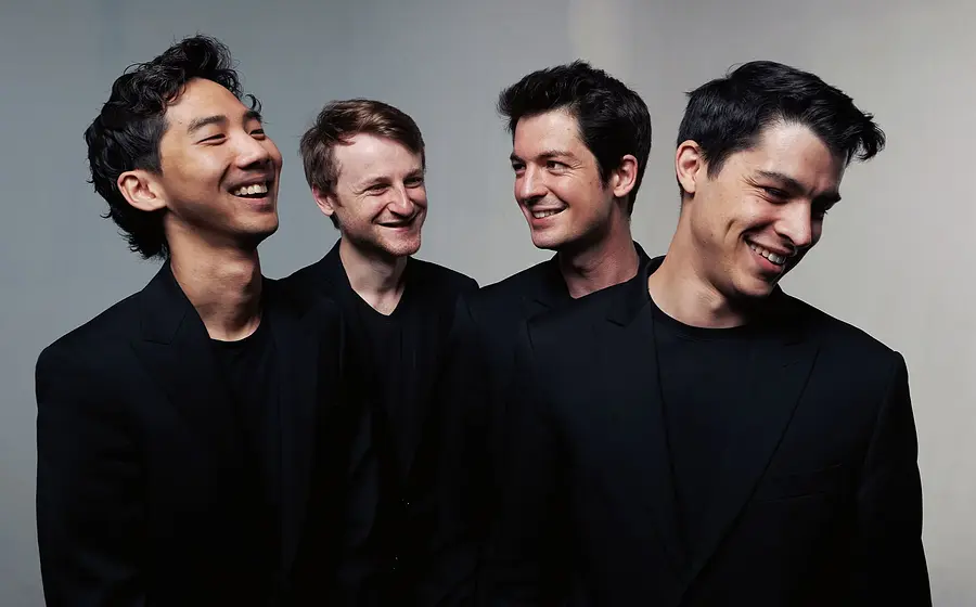 Quatuor Arod mit vier Musiker:innen in schwarzen Anzügen vor hellem Hintergrund, nebeneinander stehend und einander zugewandt, ohne Instrumente im Bild sichtbar.