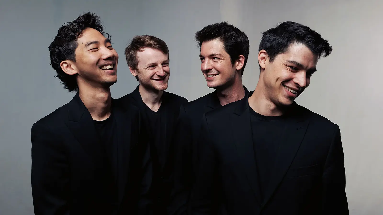 Quatuor Arod mit vier Musiker:innen in schwarzen Anzügen vor hellem Hintergrund, nebeneinander stehend und einander zugewandt, ohne Instrumente im Bild sichtbar.