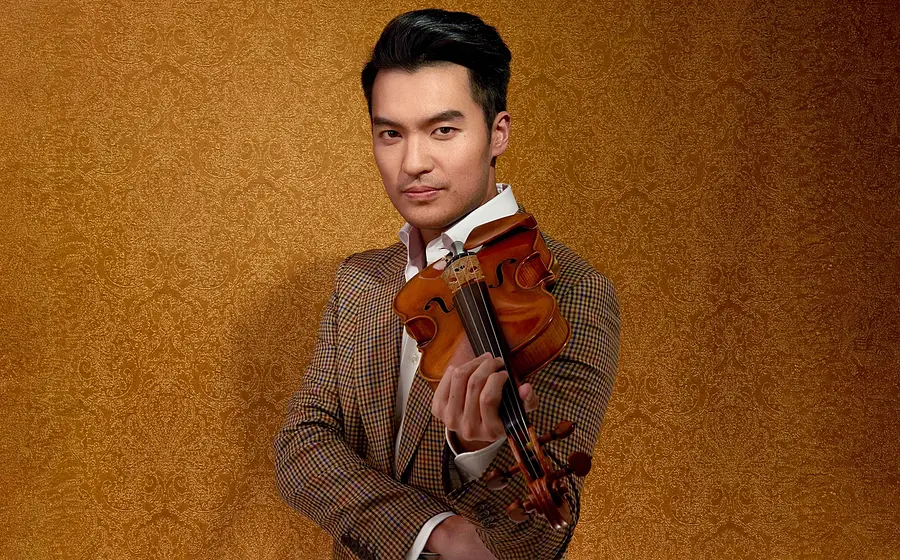 Ray Chen hält eine Violine und trägt ein gemustertes Jackett vor goldfarbenem Hintergrund.