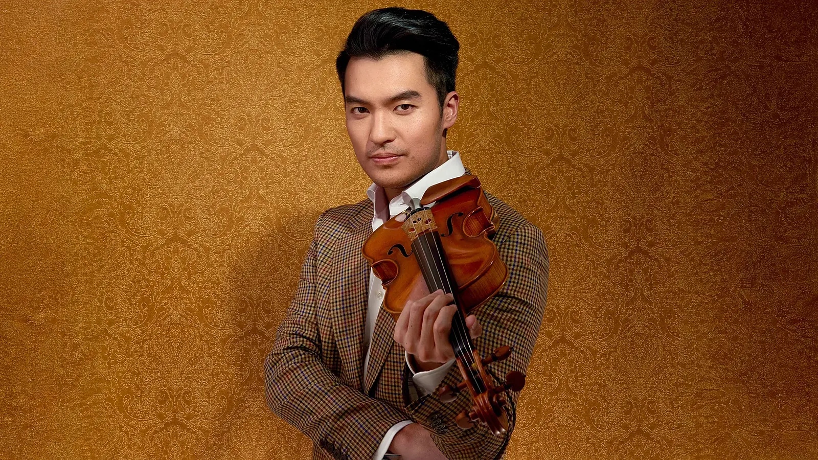 Ray Chen hält eine Violine und trägt ein gemustertes Jackett vor goldfarbenem Hintergrund.
