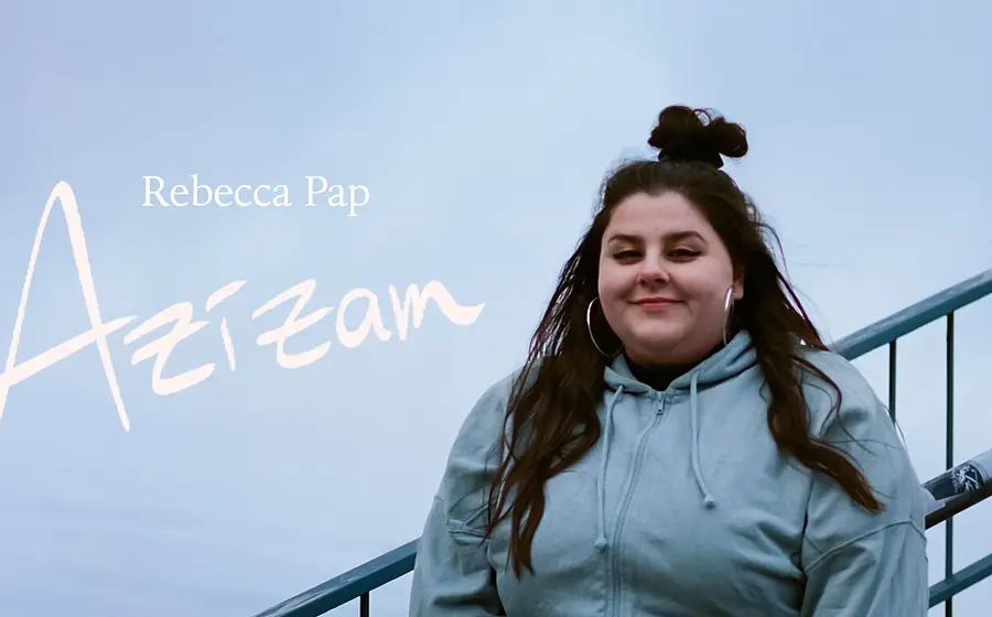 Eine Person steht draußen vor einem hellen, nebligen Hintergrund an einem Metallgeländer. Links im Bild steht der Schriftzug ›Rebecca Pap – Azizam‹