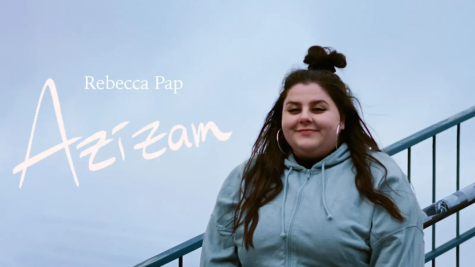 Eine Person steht draußen vor einem hellen, nebligen Hintergrund an einem Metallgeländer. Links im Bild steht der Schriftzug ›Rebecca Pap – Azizam‹