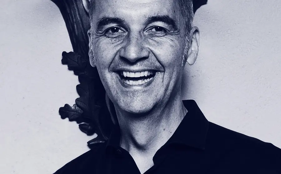 Porträt von Stefan Kröll in dunklem Hemd vor heller Wand mit dekorativem Wandelement im Hintergrund