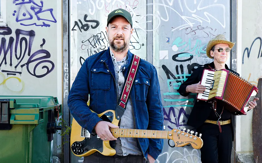 Zwei Musiker vor einer mit Graffiti besprühten Wand. Im Vordergrund steht eine Person mit E-Gitarre und Kappe, im Hintergrund spielt eine Person mit Hut und Sonnenbrille Akkordeon neben einem grünen Müllcontainer.