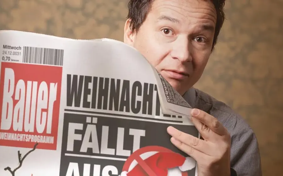 Person mit braunen Haaren und Blick in die Kamera hält eine Zeitung mit der Schlagzeile Weihnachten fällt aus Josef gesteht alles und einer durchgestrichenen Weihnachtsmütze vor sich, vor einem warm beleuchteten Hintergrund.