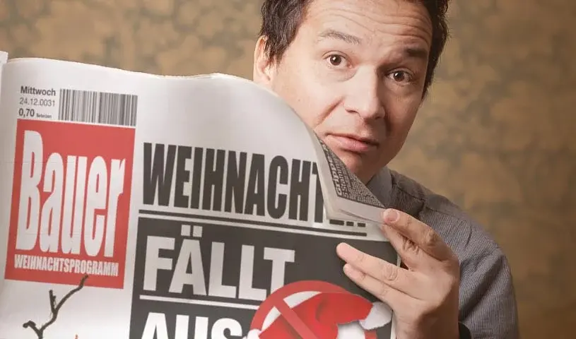 Person mit braunen Haaren und Blick in die Kamera hält eine Zeitung mit der Schlagzeile Weihnachten fällt aus Josef gesteht alles und einer durchgestrichenen Weihnachtsmütze vor sich, vor einem warm beleuchteten Hintergrund.