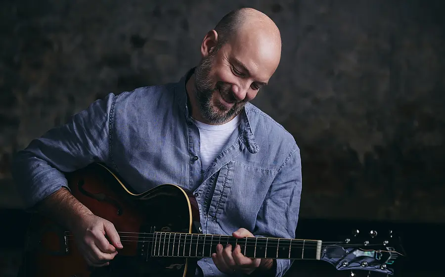 Person spielt eine E‑Gitarre vor neutralem Hintergrund, nach unten blickend, in heller Kleidung und mit Nahaufnahme der Hände auf den Saiten.