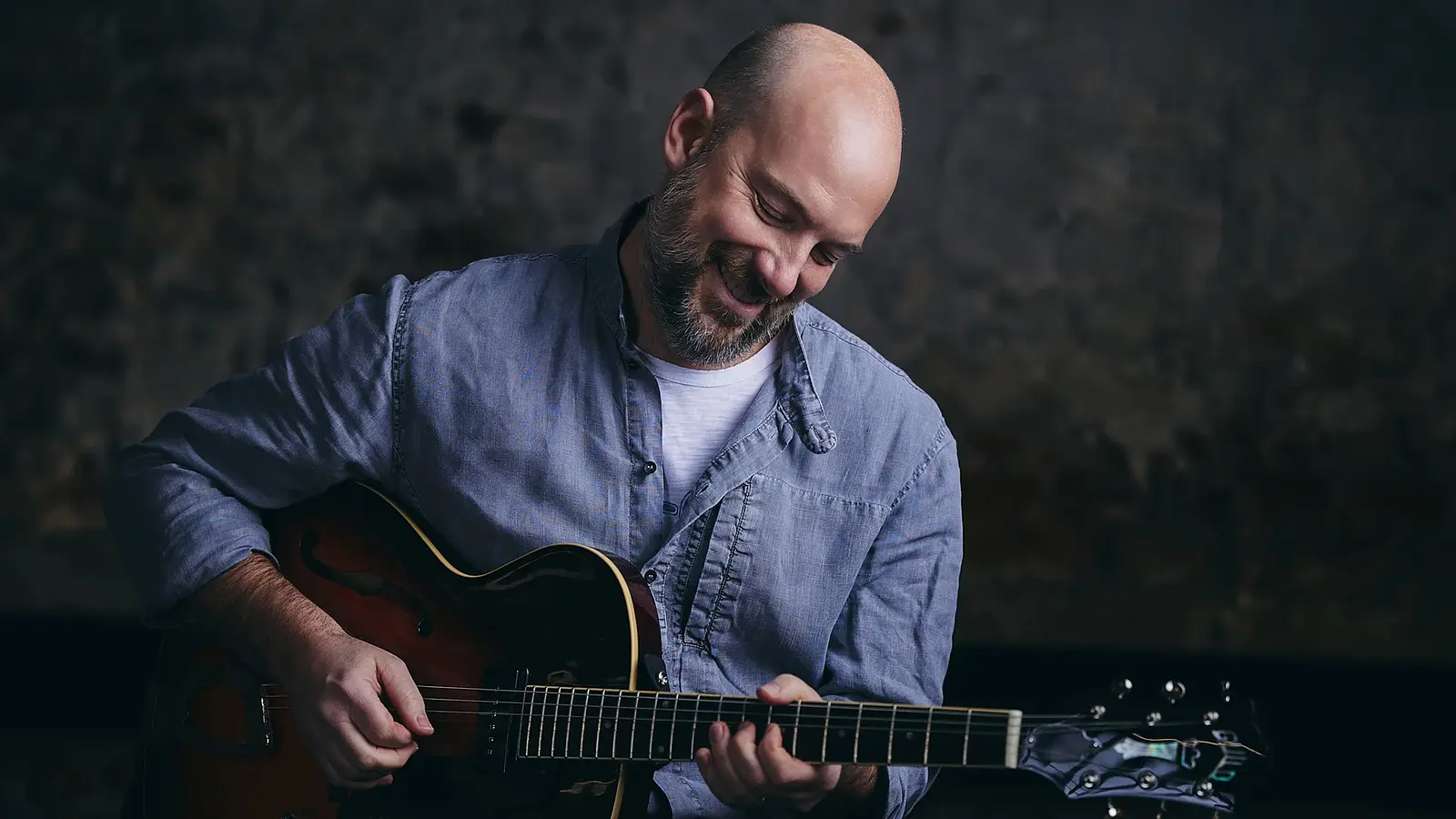 Person spielt eine E‑Gitarre vor neutralem Hintergrund, nach unten blickend, in heller Kleidung und mit Nahaufnahme der Hände auf den Saiten.