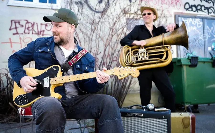 Foto zu „Stephan Zinner – Der Teufel, das Mädchen, der Blues und Ich“ mit zwei Musikern im Freien, einer mit E-Gitarre sitzend, der andere mit Tuba stehend vor einer Hauswand mit Graffiti und Mülltonnen.