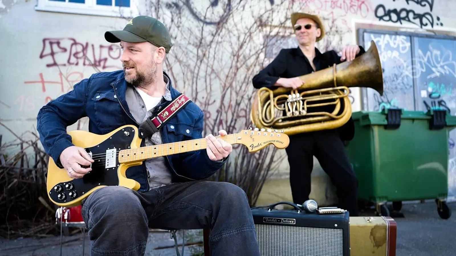 Foto zu „Stephan Zinner – Der Teufel, das Mädchen, der Blues und Ich“ mit zwei Musikern im Freien, einer mit E-Gitarre sitzend, der andere mit Tuba stehend vor einer Hauswand mit Graffiti und Mülltonnen.