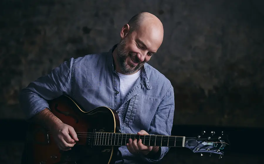 Stephan Zinner in Jeanshemd mit Gitarre vor dunkel marmoriertem Hintergrund