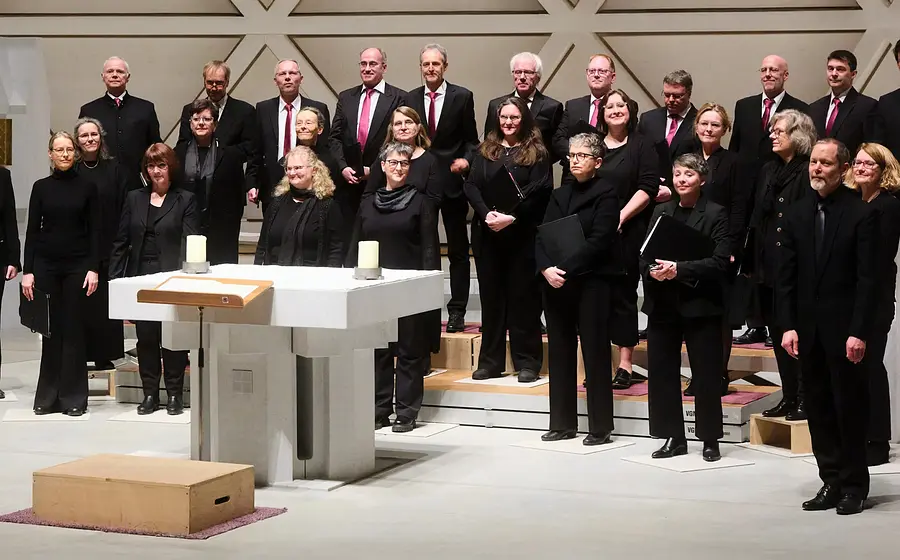 Gruppenfoto des Chors Vox Nova mit zahlreichen Chormitgliedern in schwarzer Konzertkleidung auf Podesten in einer modernen Kirche, vor einem weißen Altar mit Kerzen und geometrisch gestalteter Rückwand.