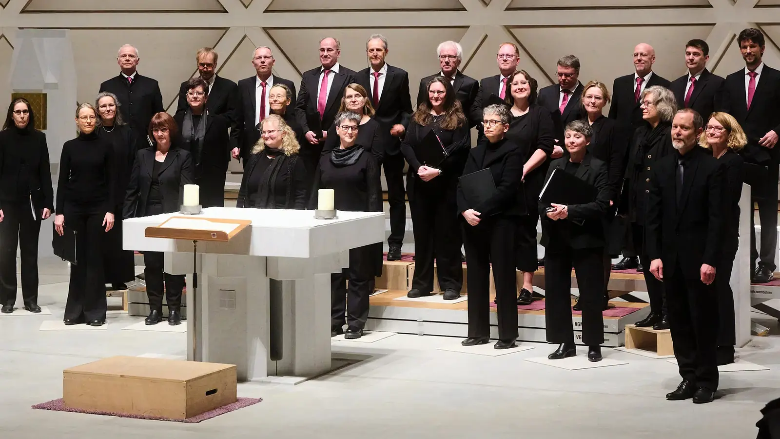Gruppenfoto des Chors Vox Nova mit zahlreichen Chormitgliedern in schwarzer Konzertkleidung auf Podesten in einer modernen Kirche, vor einem weißen Altar mit Kerzen und geometrisch gestalteter Rückwand.