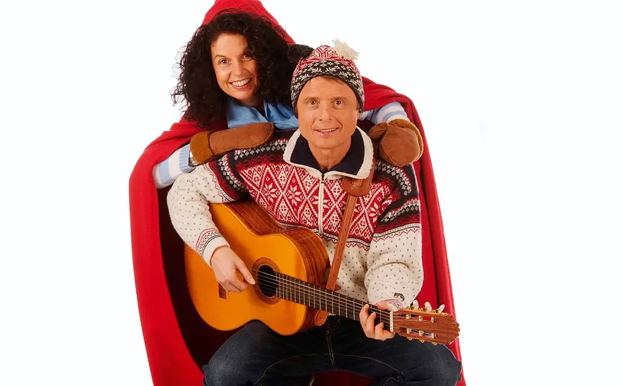 Pressefoto von Sternschnuppe mit zwei Personen in winterlicher Kleidung, eine Person mit Akustikgitarre in norwegischem Strickpullover und Mütze, die andere mit rotem Umhang und Fäustlingen, vor hellem Hintergrund