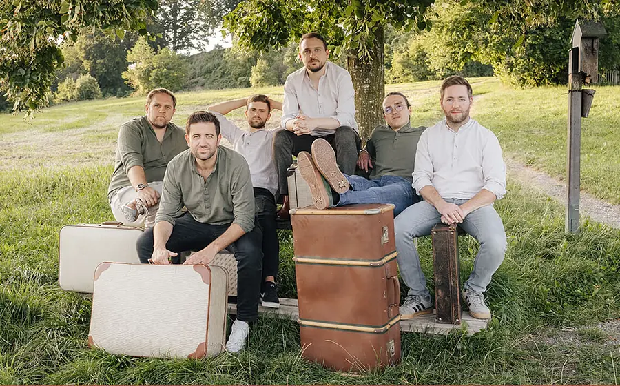 Eine Gruppe von sechs Personen sitzt entspannt auf einer Holzbank unter einem Baum im Grünen. Vor ihnen stehen mehrere große, vintage‑artige Reisekoffer. Die Szene wirkt ländlich und sommerlich, mit Wiese, Bäumen und einem Weg im Hintergrund.