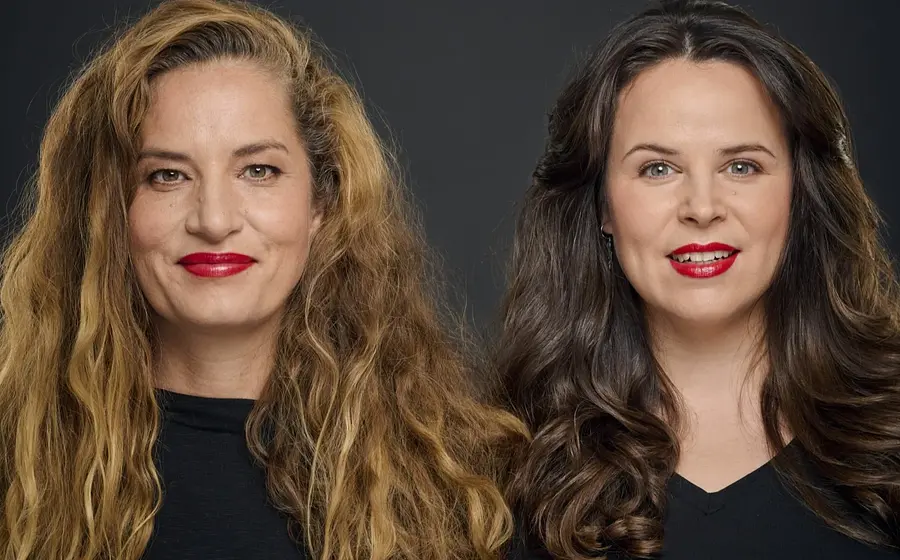 Zwei Damen mit langen Haaren und rotem Lippenstift in schwarzer Kleidung vor dunklem Hintergrund