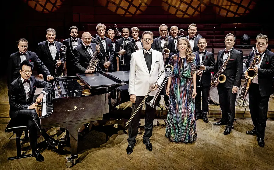 Eine Bigband in formeller Kleidung steht auf einer Konzertbühne; im Vordergrund ein Flügel sowie zwei Musiker*innen mit Posaune und Mikrofon, dahinter weitere Bläser des Ensembles.