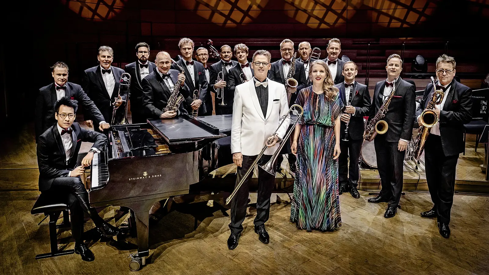 Eine Bigband in formeller Kleidung steht auf einer Konzertbühne; im Vordergrund ein Flügel sowie zwei Musiker*innen mit Posaune und Mikrofon, dahinter weitere Bläser des Ensembles.