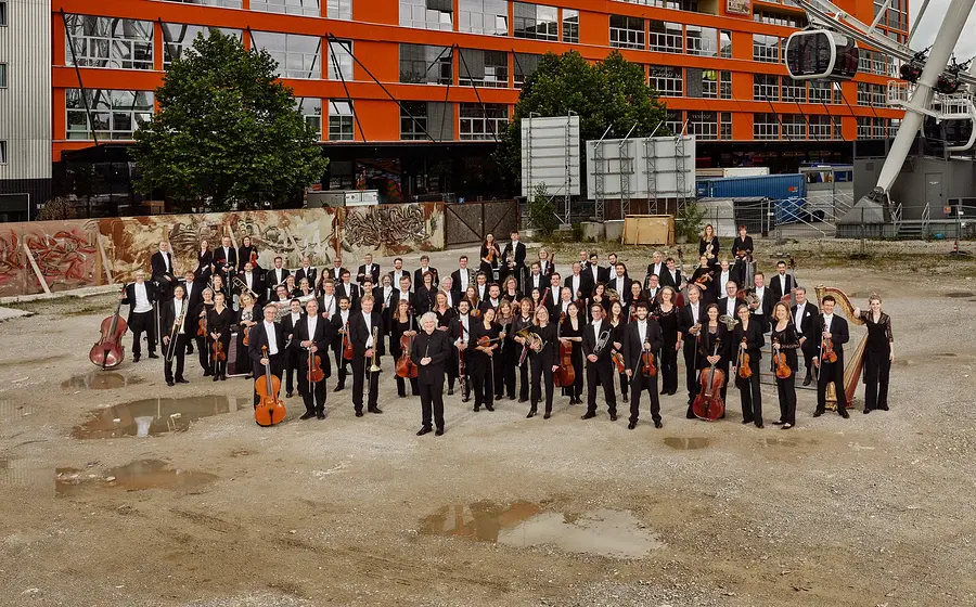 Das Orchester des Bayerischen Rundfunks, schwarz gekleidet, steht im freien auf einer ungeteerten Fläche mit Pfützen, im Hintergrund die Fassade eines großen Blocks